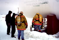 A group of people standing near an anhydrous ammonia tank in the winter. Two people are wearing fire fighter jackets