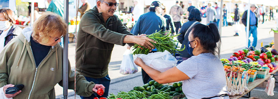 man buying vegetables from a vendor at a farmers' market and woman looking at the produce