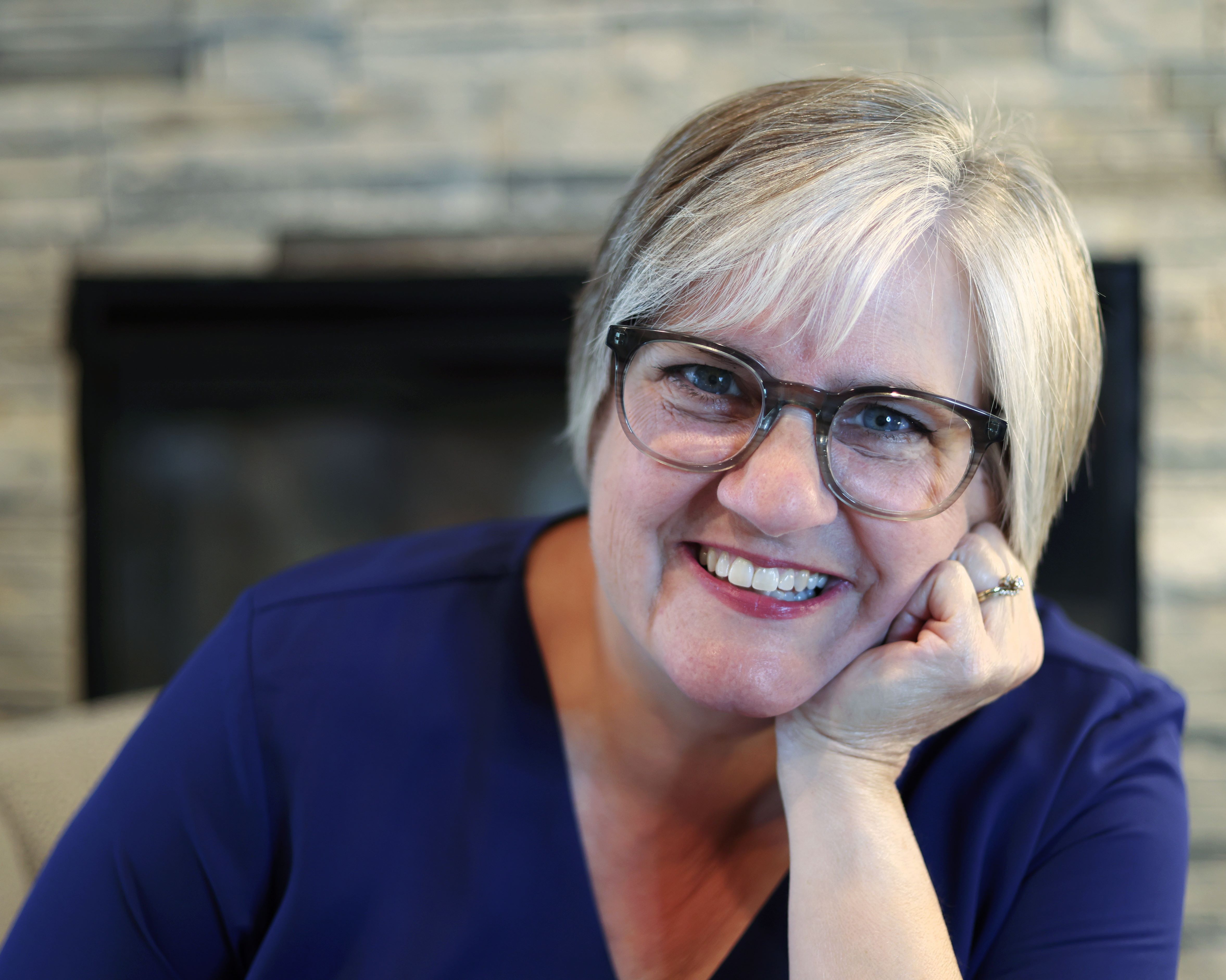Smiling woman with short blonde hair, blue eyes, and glasses wearing a royal blue shirt in front of a fireplace