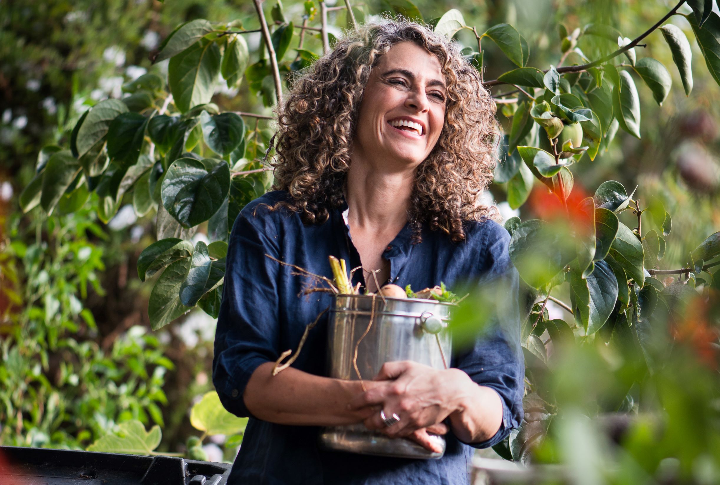 Laughing woman with shoulder length curly gray-brown hair wearing a dark blue shirt, holding a silver bucket of vegetables, and surrounded by green leaves