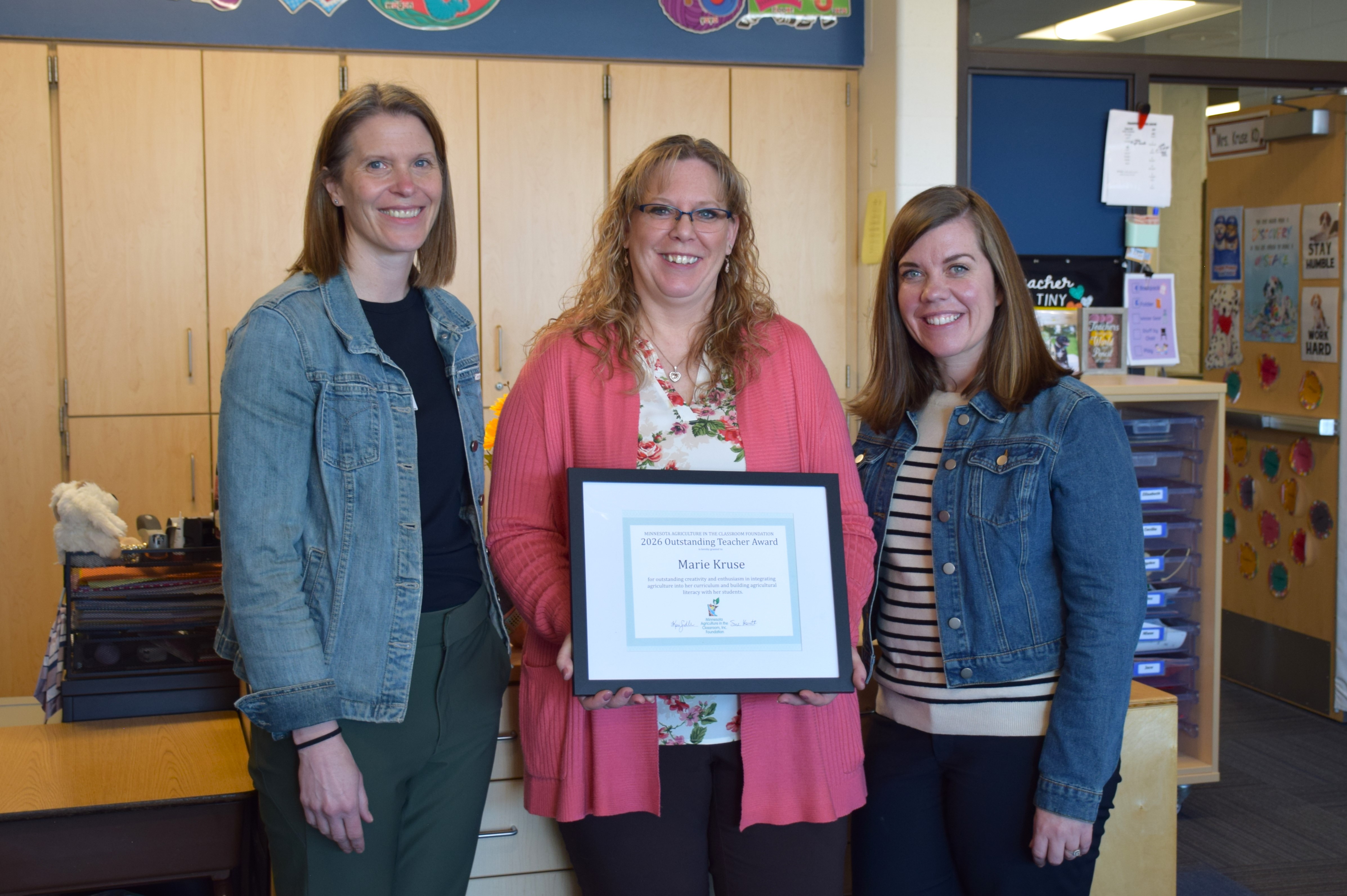 Marie Kruse holds her 2026 Outstanding Teacher Award certificate, standing with two Minnesota Ag in the Classroom team members in a classroom.